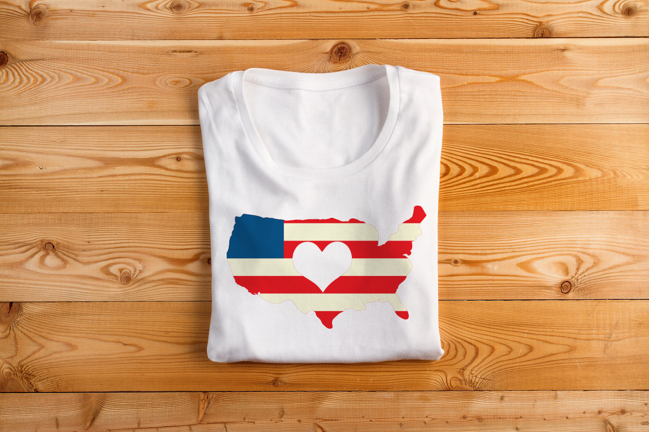 Folded shirt with image of the United states. It has a blue square in the corner and red and white stripes, like an American flag. A heart is knocked out of the middle.