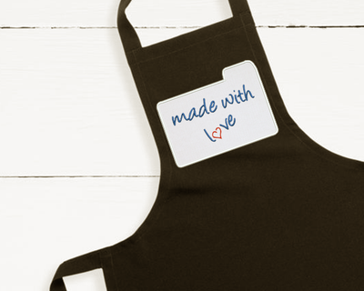 Apron with applique of recipe card that says "made with love." There is a heart in place of the O.