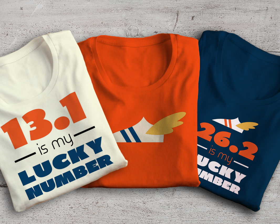 Three folded tees. One says "13.1 is my lucky number," one has a winged sneaker, and one says "26.2 is my lucky number" with a winged sneaker above.