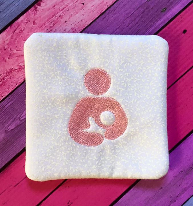 A square white fabric coaster sits on a pink and purple stained wood background. Embroidered onto the coaster in pink is the breastfeeding icon.