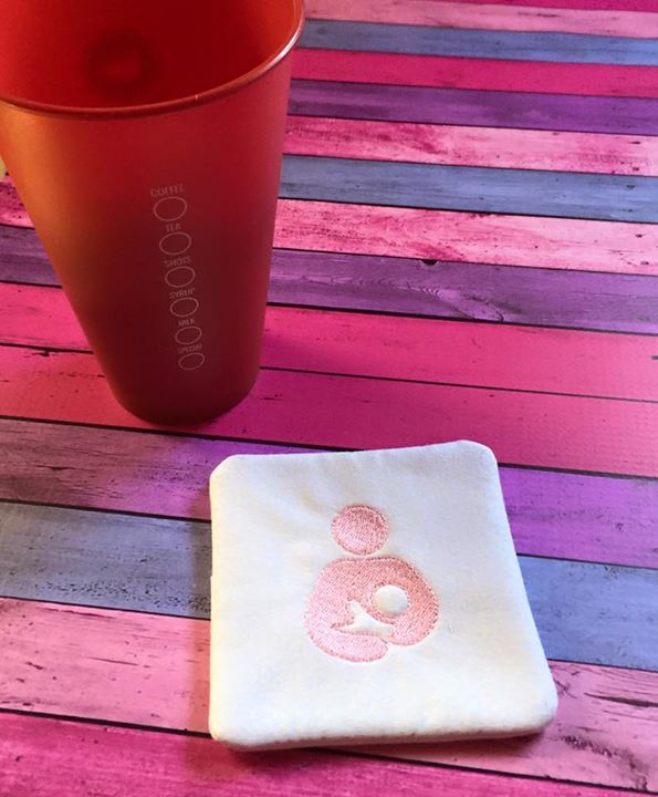 A square white fabric coaster sits on a pink and purple stained wood background. Embroidered onto the coaster in pink is the breastfeeding icon. A red cup sits nearby.