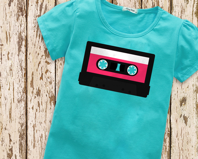 A turquoise child's shirt lays on a weathered painted wood background. The shirt has a cassette tape design with a hot pink label.