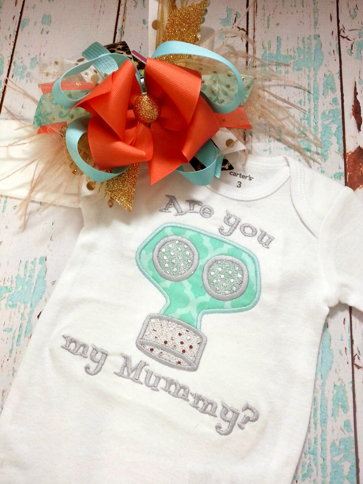 A white baby sleeper sits on a weathered painted wood background. Above the sleeper is a gold, coral, and aqua bow. The shirt is embroidered with the phrase "Are you my Mummy?" and a gas mask done in aqua quatrefoil and silver fabric.