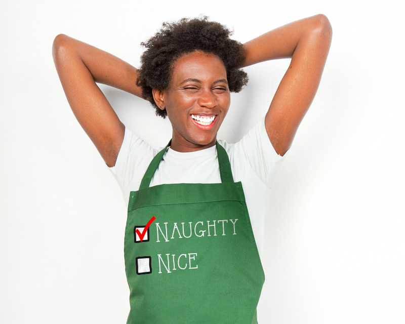 Black woman smiling and wearing an apron that says "Naughty" and "Nice" and naughty is checked off.