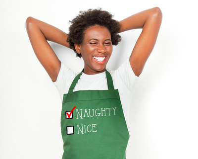 Black woman smiling and wearing an apron that says "Naughty" and "Nice" and naughty is checked off.