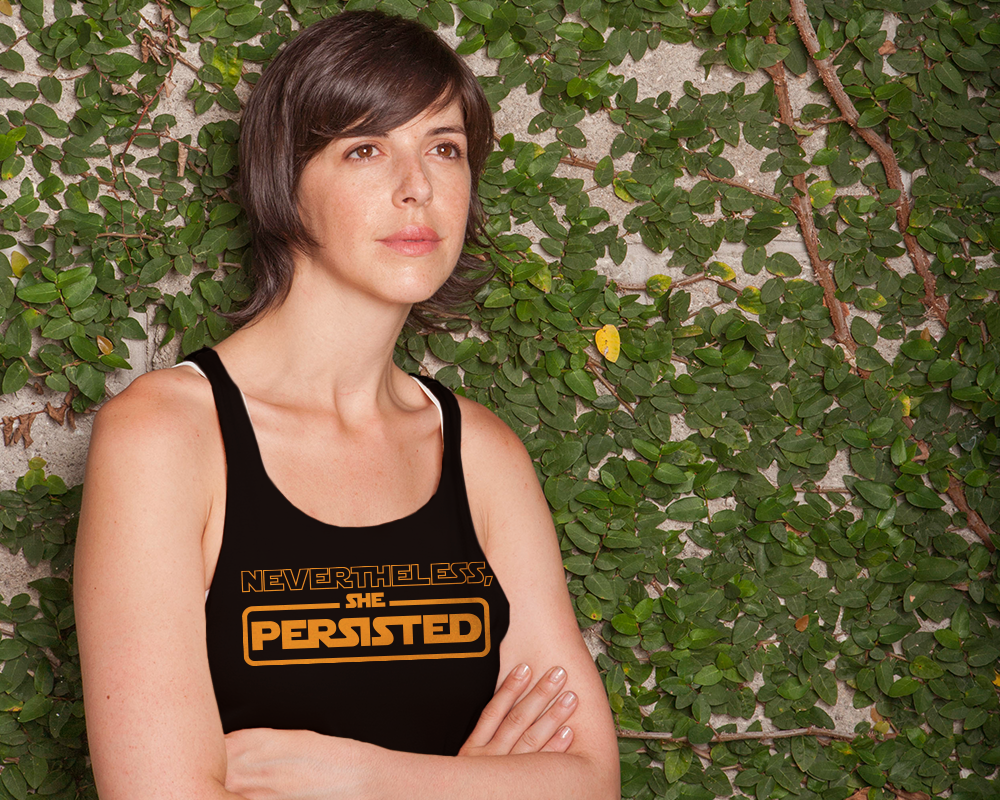 White-presenting Latinx woman with arms crossed. She wears a tank top that says "nevertheless she persisted."
