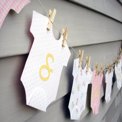 Paper bodysuits in different pastel patterns and colors hanging from a clothesline with paper letters glued on top.