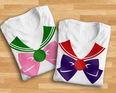 Two folded white tees with a sailor collar, large bow, and medallion design. The left shirt has a pink and green design, the right shirt has a purple and red design.