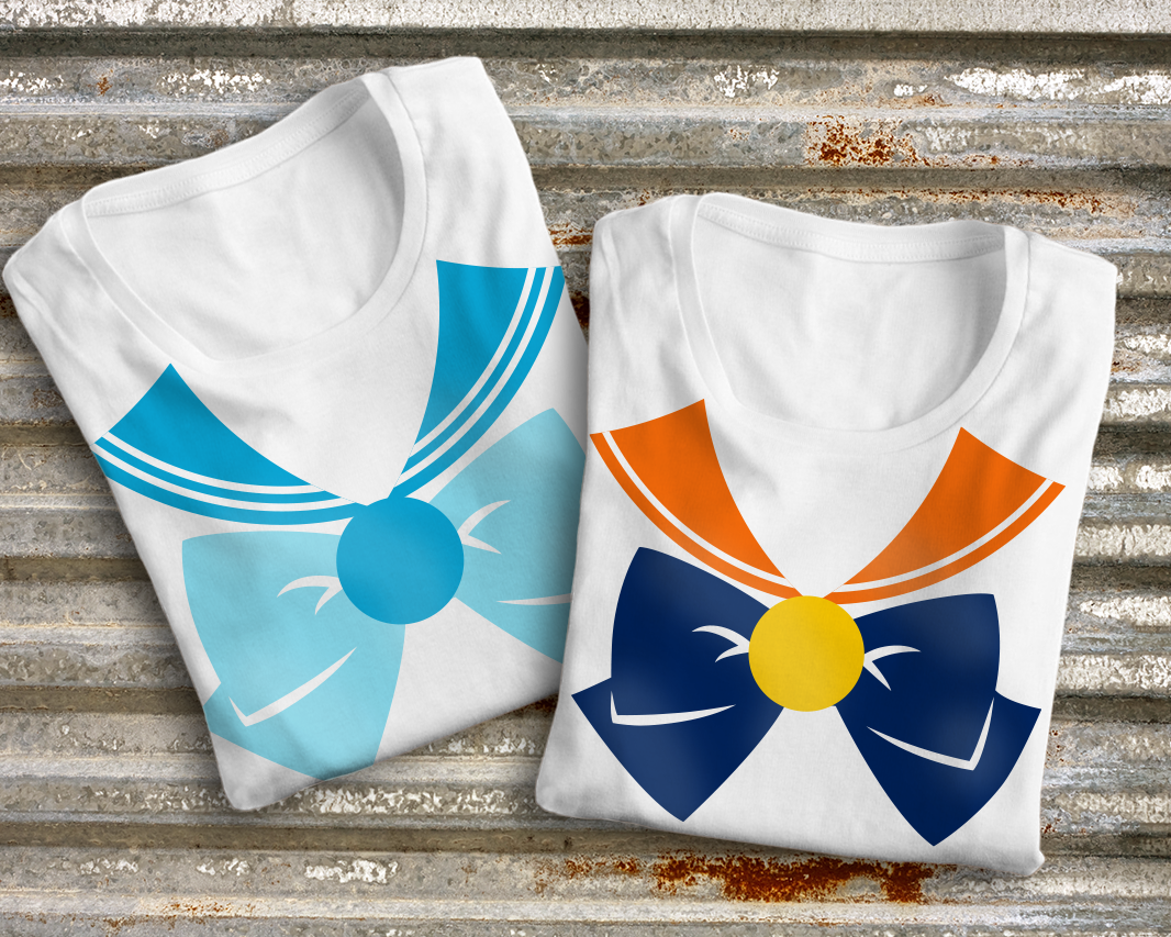 Two folded white shirts with sailor collars, large bows, and medallions. The left shirt has the design in shades of blue, the right shirt has an orange, blue, and yellow design.