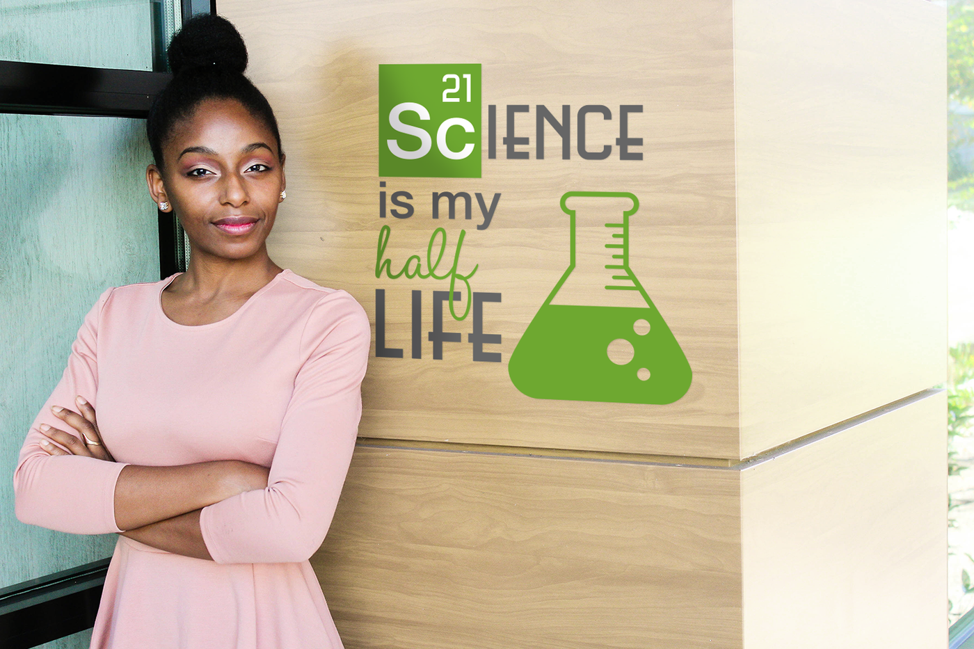 Black woman stands in front of a wall with a smile and folded arms. On the wall is a design that says "Science is my half life" with a scientific flask.