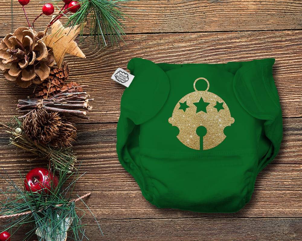 A green cloth diaper has a gold glitter round bell design. It is on a wood background with festive Christmas greenery to the side.