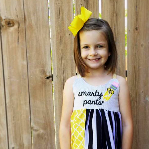 White little girl wears a dress with an applique pencil wearing glasses and the embroidered phrase "smarty pants."
