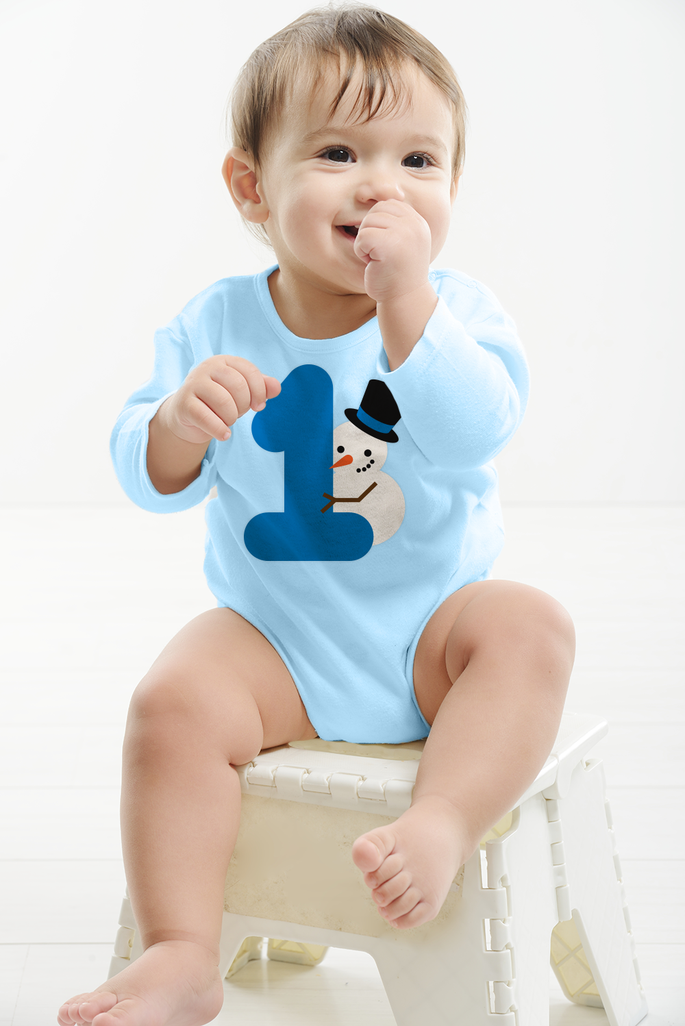 A white baby sucks his thumb. He has a blue onesie with a large 1 and a snowman behind the number.