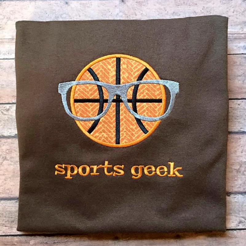 A brown square piece of fabric sits on a wooden surface. Embroidered onto the fabric are the words "sports geek" and an applique basketball wearing geeky glasses.