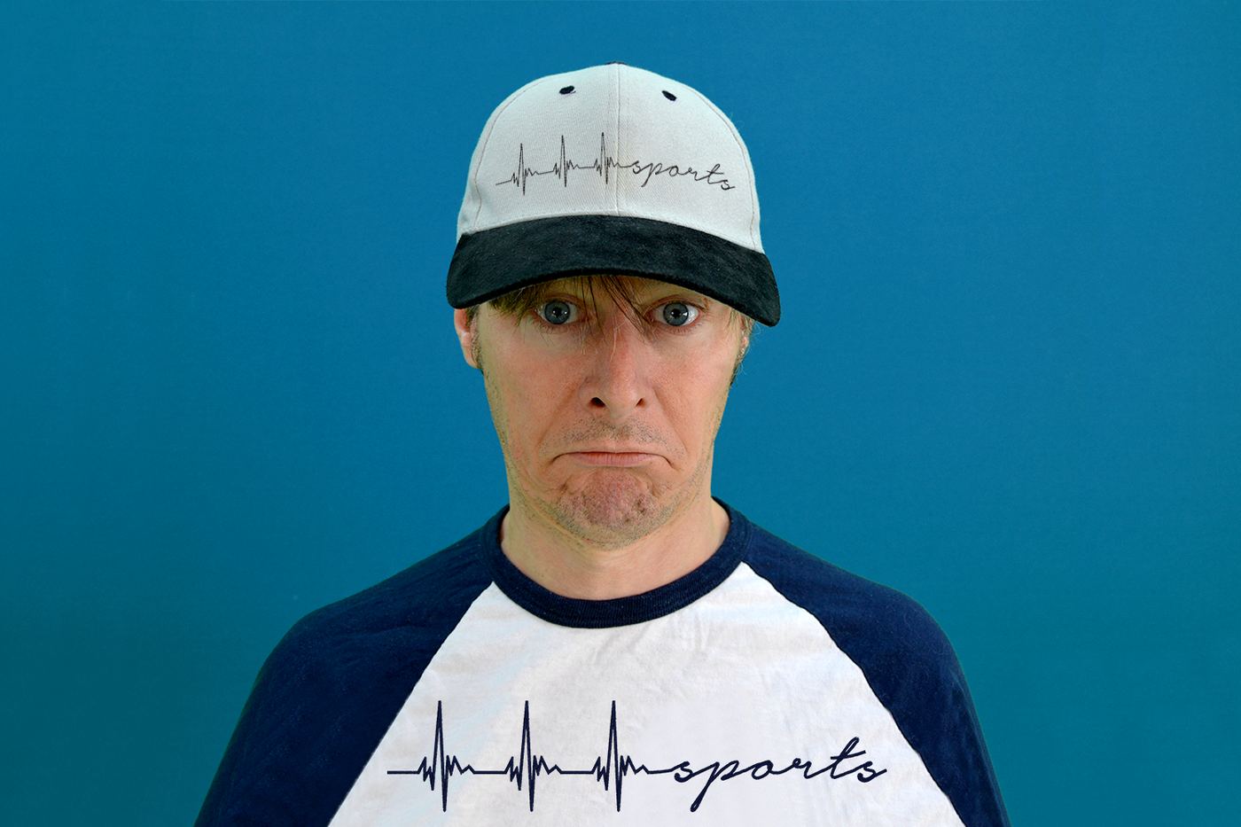 White man wearing a baseball cap and raglan tee. Both items of clothing have a heartbeat embroidery that ends in the word "sports"