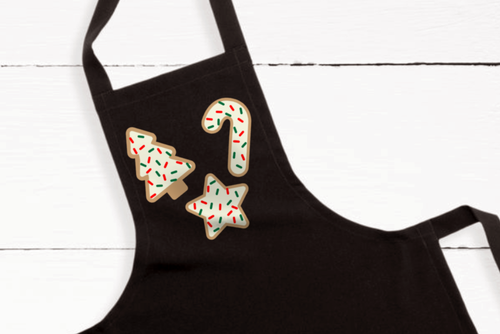 A black apron that has the image of 3 frosted sugar cookies with red and green sprinkles. The sugar cookies are in the shape of a tree, a star, and a candy cane.