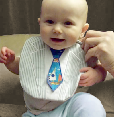 Smiling white baby  is wearing a bib that looks like a suit, complete with a neck tie.