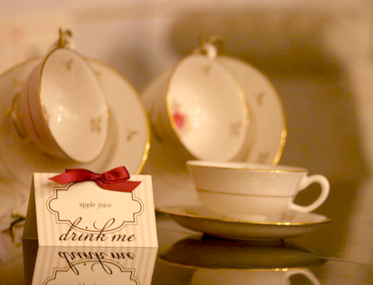 Three tea cups with a table tent in front. The tent is ivory with tan stripes and a label that says "apple juice. " Below it says "drink me."