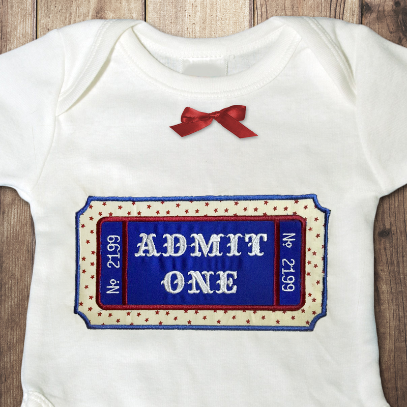 A white baby onesie with a red bow at the top. Embroidered onto the onesie is a large carnival ticket. The center area is blue applique and outside there is pale yellow applique fabric with stars. The text on the ticket says "ADMIT ONE" and "No. 2199" on each side.