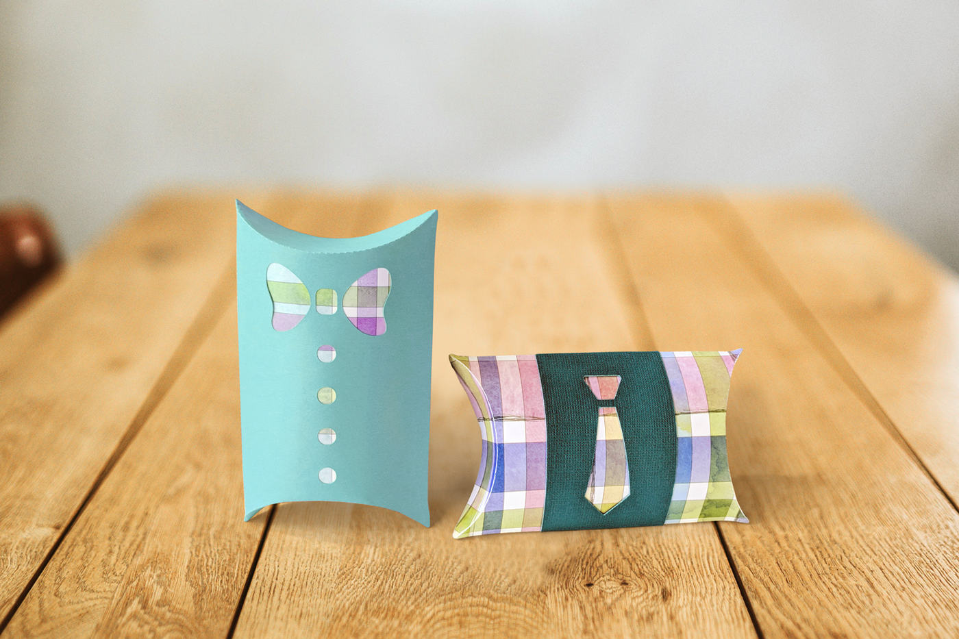 Two pillow boxes on a wood table. The left box has a bowtie and buttons cutout revealing plaid paper underneath. The right box is plaid with a band that has a necktie cutout that shows the pattern of the box through the hole.