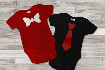A red and black onesie on a wood background. One has a white bow tie, the other has a red neck tie.