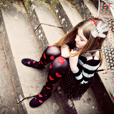A white girl holding an umbrella sits on some steps. She wears a red, black and white outfit, and a red headband with a large white cartoon glove on it.