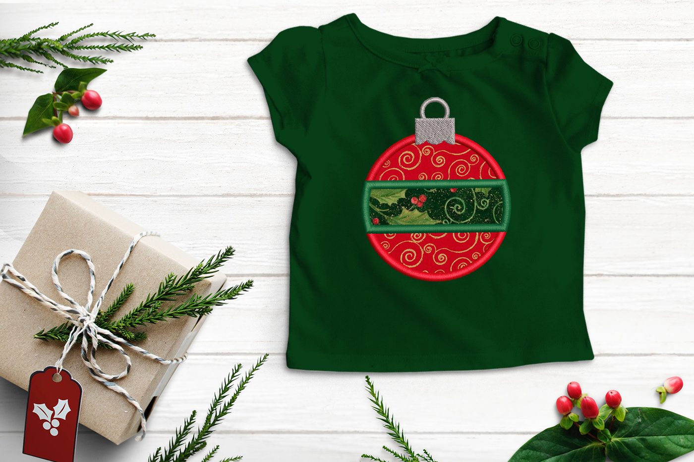 A green child's shirt sits on a white surface with a present and evergreen sprigs around it. Embroidered on the shirt is a round applique ornament with a contrasting center band. The applique fabric is red with gold swirls on the top and bottom and the center band applique is green with holly.