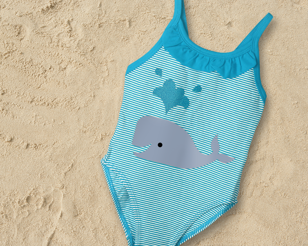 A blue and white striped child's swimsuit on some sand. A whale with water coming out of its spout decorates it.