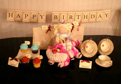 A party banner made of playing cards that says "Happy Birthday." In front on a table are Alice in Wonderland plush toys, cupcakes, party bags, and tea cups.